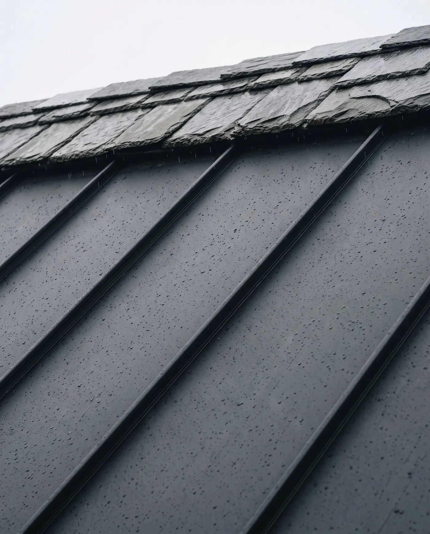 Slate tiles above standing-seam steel panels, wet with rain, seen from below.