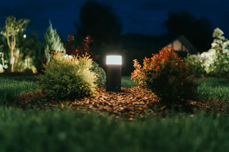 A low bollard light among shrubs and bark mulch in a garden at blue hour.